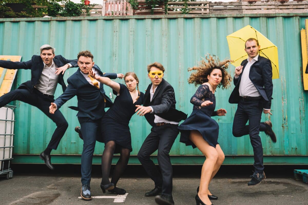 Die Affirmative - Sechs Personen springen vor einem grünen Container in die Luft